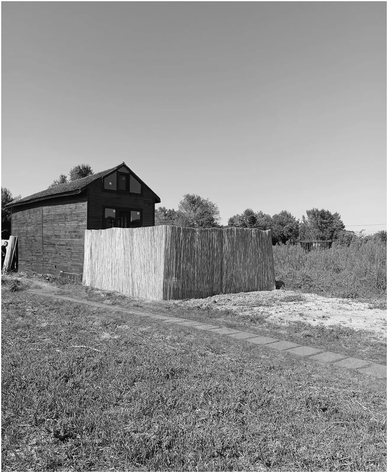 Dark timber house behind a reed fence, with a meadow and path in front; black and white.