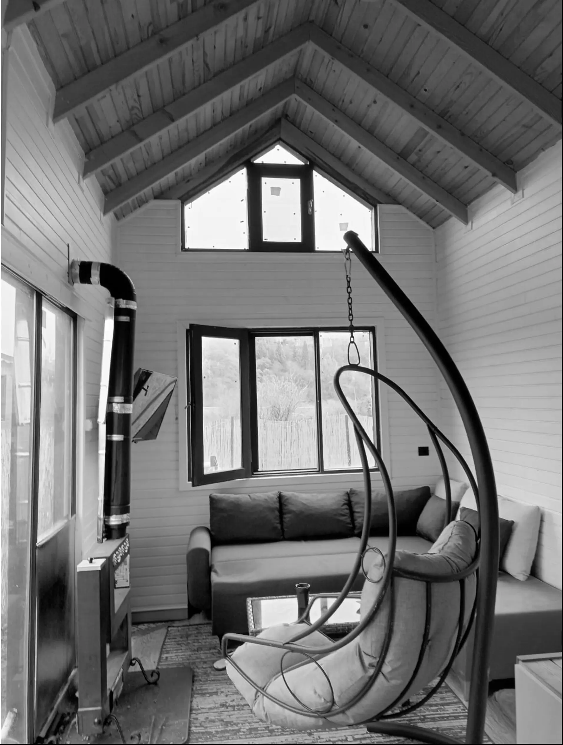 Living area with a hanging swing chair, sofa, stove flue and gable window; black and white.