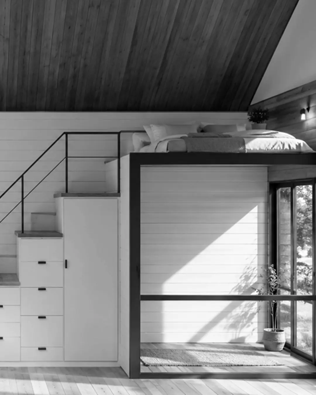 Mezzanine bed reached by a storage stair, white boarded walls and full-height glazing; black and white.