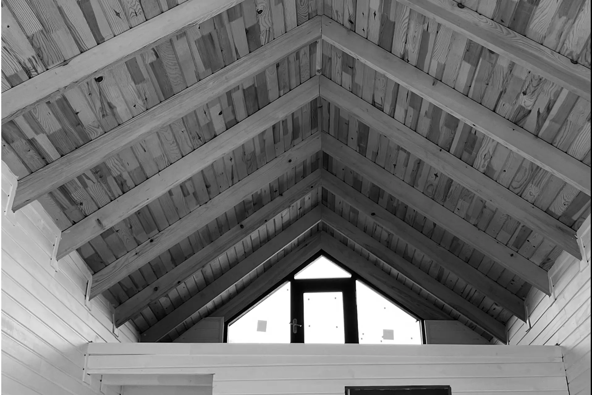 Underside of the timber gable roof: rafters and boarded ceiling converging towards a triangular window; black and white.