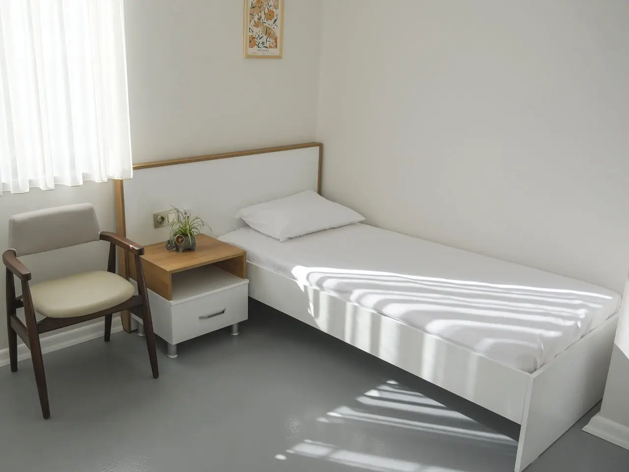 Single room with a white bed, an armchair and sunlight patterns on the floor.