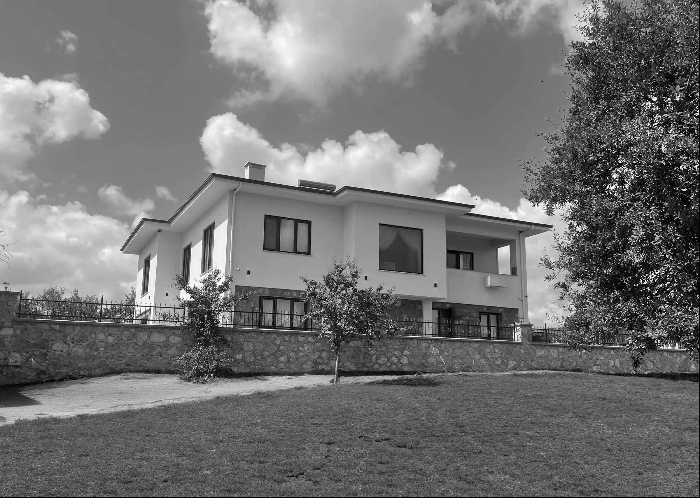 Bahçeden ev: çim, taş bahçe duvarı ve ağaçlar; bulutlu gökyüzü; siyah-beyaz.